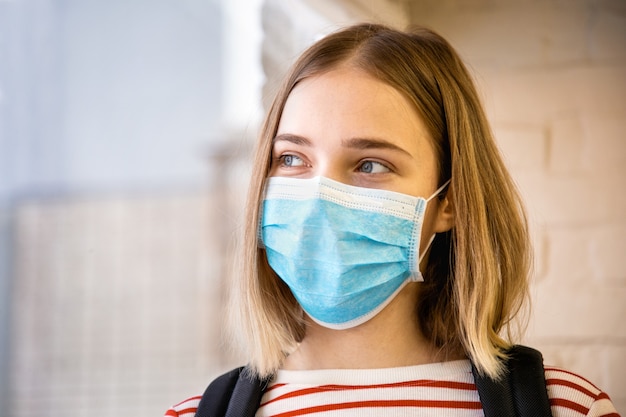Retrato de estudiante rubia junto a la ventana en máscara azul desechable. en la universidad durante el encierro del covid por coronavirus. Estudiante en máscara médica protectora.