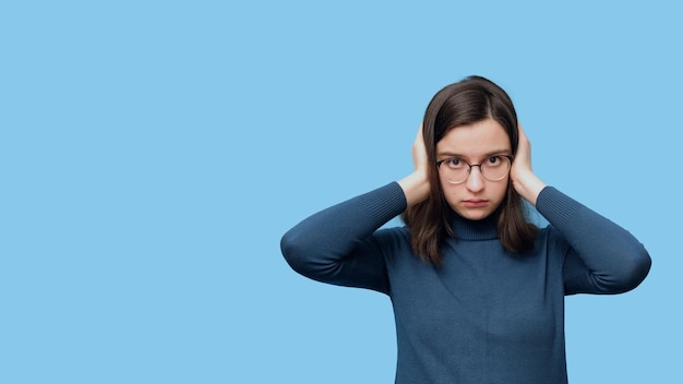Retrato de una estudiante seria con gafas y un suéter que cubre sus oídos con las manos aisladamente en un azul