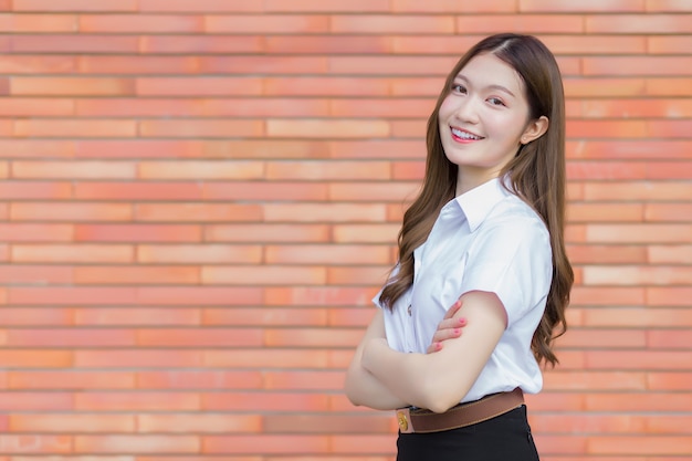 Retrato de un estudiante tailandés adulto en uniforme de estudiante universitario. Hermosa chica asiática de pie con los brazos cruzados sobre un fondo de ladrillo.