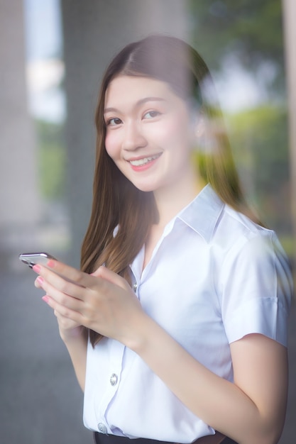 Retrato de un estudiante tailandés adulto en uniforme universitario. Hermosa mujer asiática de pie cerca de la ventana sonriendo felizmente usa su teléfono inteligente para buscar información educativa.