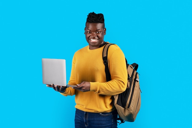 Retrato de estudiante varón negro con mochila y portátil sobre fondo azul.