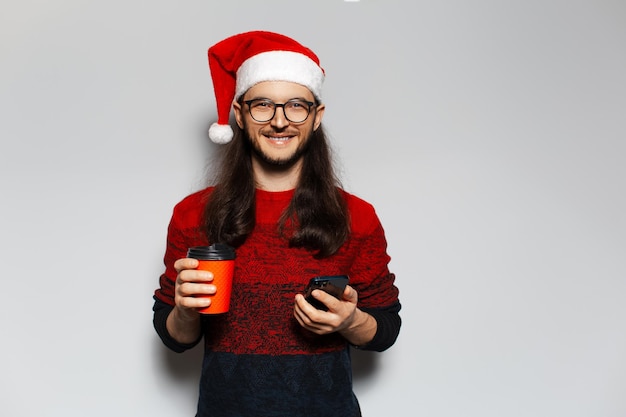 Retrato de estudio de un apuesto hombre sonriente sosteniendo un vaso de papel con una bebida para llevar y un teléfono inteligente en las manos Con sombrero de Santa Claus Concepto de Navidad Fondo blanco