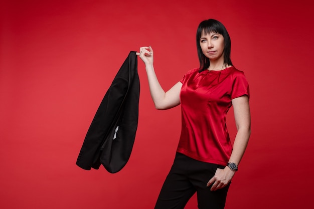 Foto retrato de estudio de atractiva empresaria sonriente vistiendo elegante traje negro con pantalones y zapatos oxford. ella sostiene los brazos cruzados sobre fondo rojo.