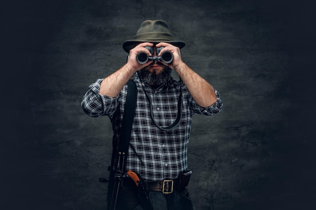 Retrato de estudio de un cazador barbudo que usa una camisa de lana a cuadros mirando a través de binoculares, sostiene un rifle.