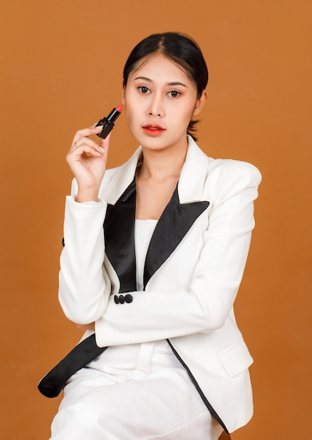 Retrato de estudio de cerca disparo modelo de mujer milenaria asiática de pelo corto cola de caballo negro usa maquillaje en blanco y negro chaqueta casual de moda brazo cruzado sosteniendo lápiz labial rojo en la mano sobre fondo marrón.