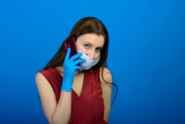 Retrato del estudio de la chica joven que lleva la máscara médica de la gripe y los guantes disponibles, presentación femenina aislada sobre fondo azul y rojo.