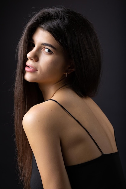 Retrato de estudio clásico de una joven morena vestida con un top negro que está sentada en una silla contra un fondo negro