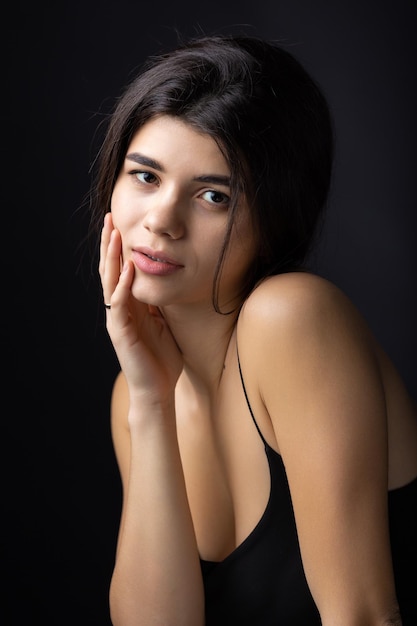 Retrato de estudio clásico de una joven morena vestida con un top negro que está sentada en una silla contra un fondo negro