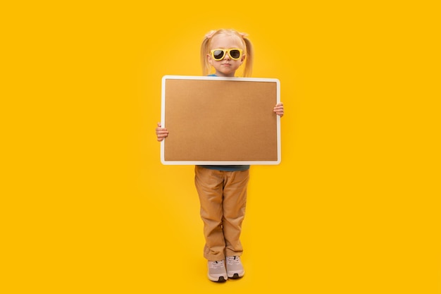 Retrato de estudio completo de una chica con gafas de sol sosteniendo un tablero de corcho vacío Espacio para tu texto Aislado en fondo amarillo