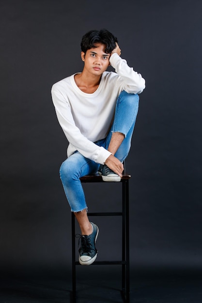 Retrato de estudio de cuerpo completo del joven asiático LGBTQ gay glamour guapo modelo masculino en camisa blanca casual jeans zapatillas sentado en una silla alta riendo sosteniendo la boca de la tapa de la mano sobre fondo negro