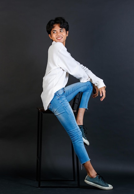 Retrato de estudio de cuerpo completo del joven asiático LGBTQ gay glamour guapo modelo masculino en camisa blanca casual jeans zapatillas sentado en una silla alta riendo sosteniendo la boca de la tapa de la mano sobre fondo negro
