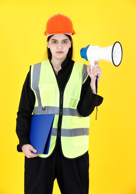 Retrato de estudio de disparo Capataz de ingeniería profesional femenina asiática con tirantes en casco duro y reflejan chaleco de seguridad sonriendo mirada a la cámara con megáfono y carpeta de documentos sobre fondo amarillo.