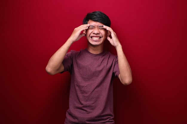 Un retrato de estudio de un doloroso joven asiático sosteniendo su cabeza sobre fondo rojo.