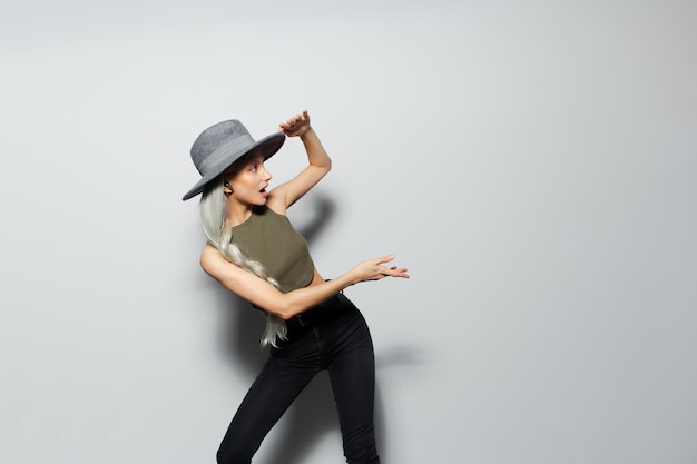 Retrato de estudio de felicidad linda chica rubia con auriculares inalámbricos en los oídos sorprendida mostrando con la mano en el espacio de copia con sombrero gris sobre fondo blanco