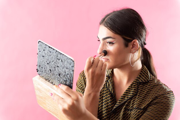 Retrato de estudio con fondo rosa de un joven que se maquilla mientras se mira en el espejo