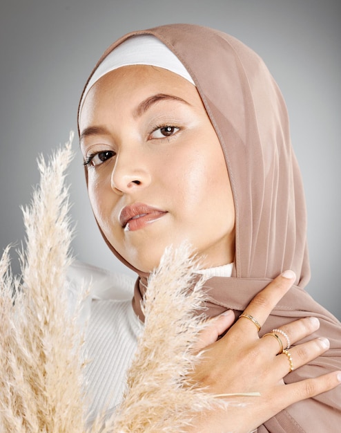 Retrato de estudio de una hermosa joven musulmana con un pañuelo marrón en la cabeza posando con una planta de trigo de las pampas contra un fondo gris Modesto árabe con maquillaje natural y cubierto con el hiyab tradicional