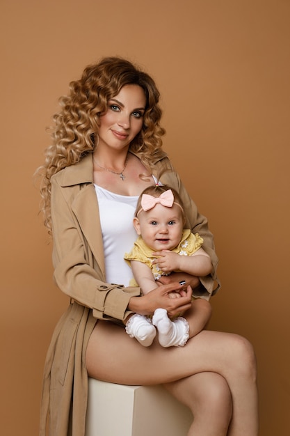 Retrato de estudio de hermosa madre caucásica con pelo largo y ondulado con trinchera clásica beige y top blanco con linda hija en brazos sentada en blanco contra la pared de melocotón.
