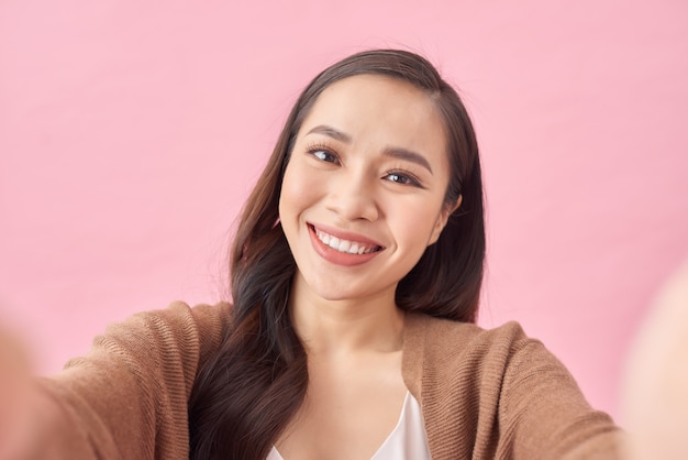 Retrato de estudio de hermosa mujer sonriendo con dientes blancos y haciendo selfie, fotografiándose a sí misma sobre fondo rosa