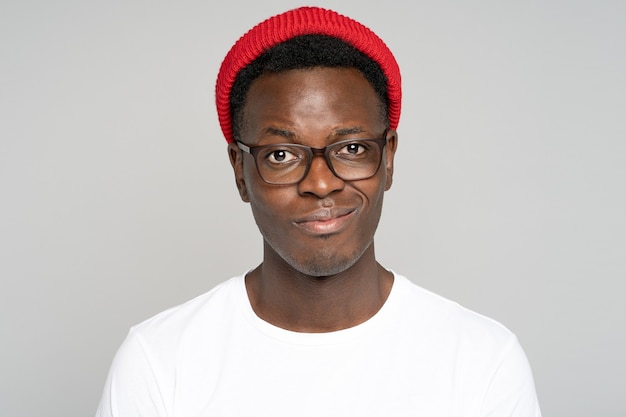 Retrato de estudio de hipster afro escéptico con gafas con sonrisa tristemente falsa, mirando con pesar a la cámara, fondo gris aislado. El negro molesto trata de mantener una actitud positiva después de fallar en algo.