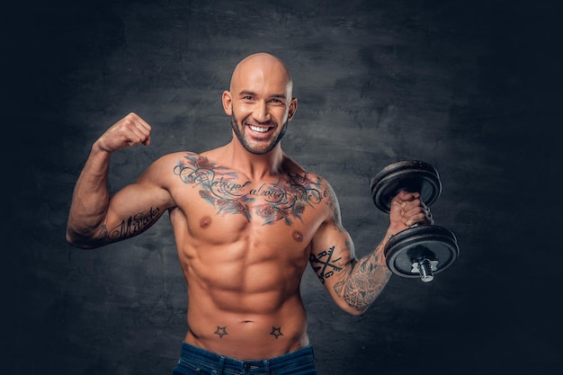 Retrato de estudio de un hombre deportivo de cabeza rapada con tatuajes en el pecho y los brazos sostiene pesas.