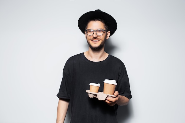 Retrato de estudio de un hombre guapo sonriente con sombrero negro con tazas de café ecológicas