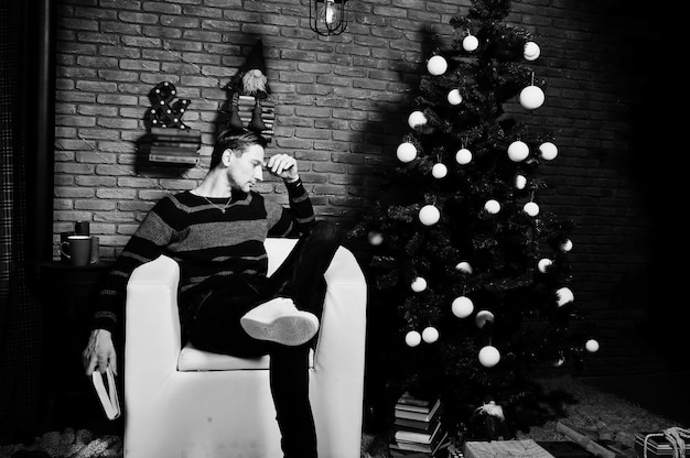 Retrato de estudio de hombre con libro sentado en una silla contra el árbol de Navidad con adornos.