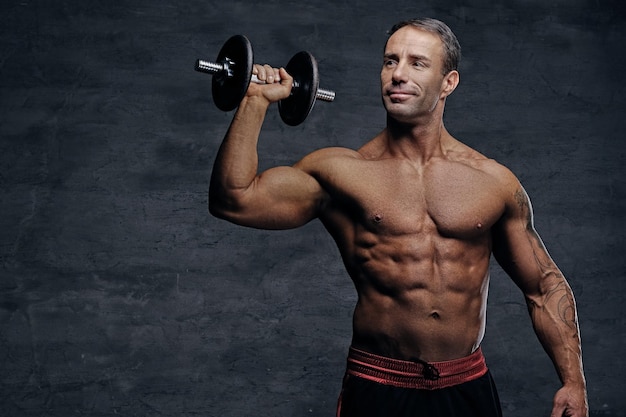 Retrato de estudio de hombre musculoso sin camisa de mediana edad sostiene dumbbel aislado sobre fondo gris.