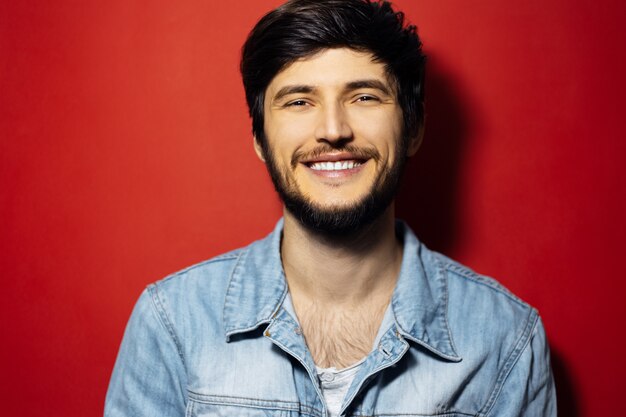 Retrato de estudio de hombre sonriente joven con chaqueta vaquera sobre superficie roja.