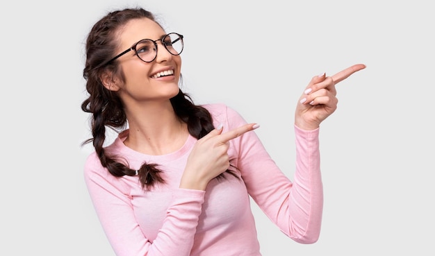 Retrato de estudio horizontal de una alegre joven morena con blusa rosa y anteojos indica a un lado con los dedos índices al espacio en blanco para su publicidad aislada sobre fondo blanco