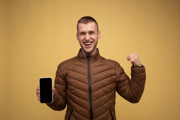 Retrato de estudio. Joven de 20 años con una chaqueta marrón, de fondo amarillo, sosteniendo en la mano, usa un teléfono móvil, hace un gesto ganador, apretando el puño, dice "sí"