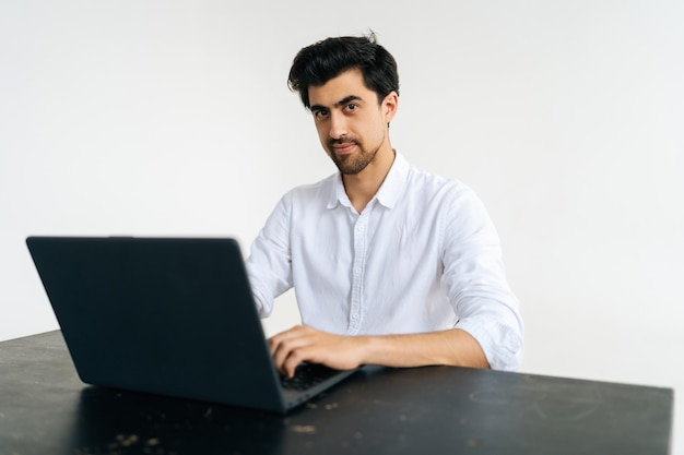 Retrato de estudio de un joven amigable con camisa que trabaja en una computadora portátil escribiendo en el teclado mirando a la cámara sentado en el escritorio en un fondo blanco aislado