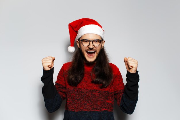 Retrato de estudio de un joven apuesto hombre feliz con el pelo largo que celebra su triunfo haciendo un gesto ganador con ambas manos lleva un suéter rojo de Navidad y un sombrero de Papá Noel en un fondo blanco