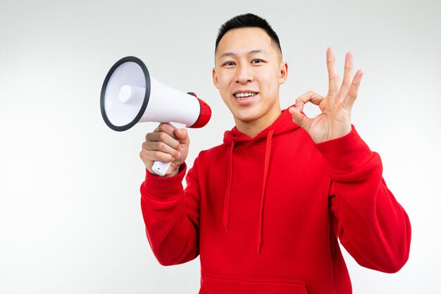 Retrato de estudio de un joven asiático en una sudadera con capucha roja con un altavoz en sus manos sobre un estudio blanco