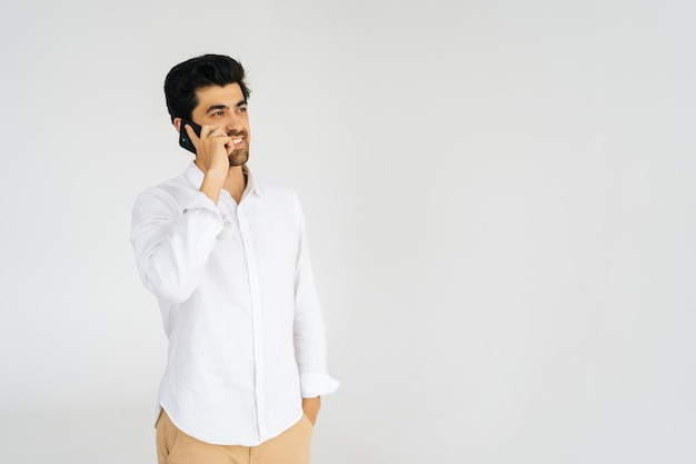 Retrato de estudio de un joven barbudo sonriente hablando de un teléfono inteligente conversando en un teléfono móvil sosteniendo y usando un teléfono celular en un fondo blanco aislado