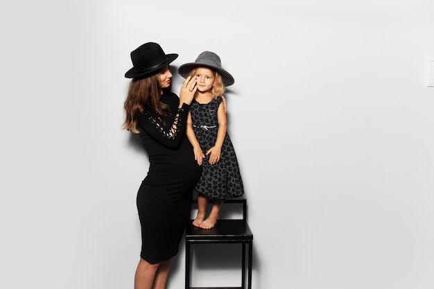 Retrato de estudio de una joven embarazada con su hija sobre fondo blanco con sombreros vestidos de negro
