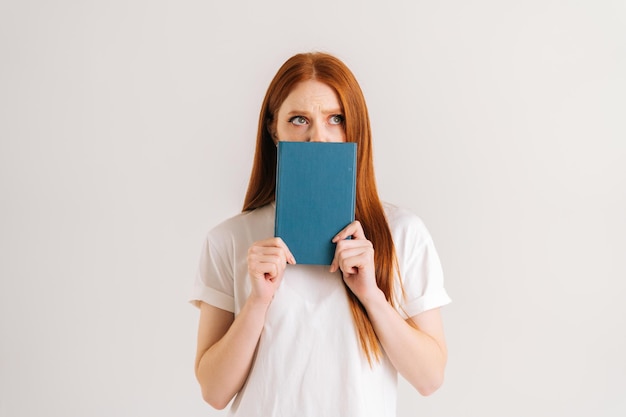 Retrato de estudio de una joven estudiante frustrada que esconde la cara detrás de un cuaderno mirando a la cámara