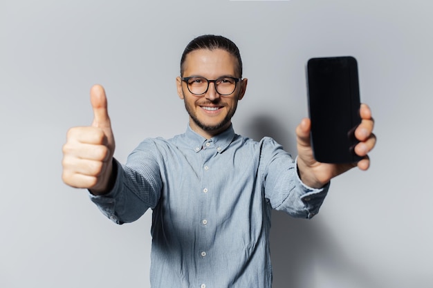 Retrato de estudio de un joven feliz que muestra un teléfono inteligente y pulgares hacia arriba sobre fondo blanco con anteojos y camisa azul