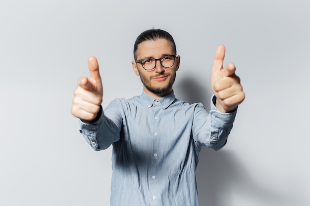 Retrato de estudio de un joven feliz que te señala con el dedo a la cámara en un fondo de color blanco con camisa azul y anteojos