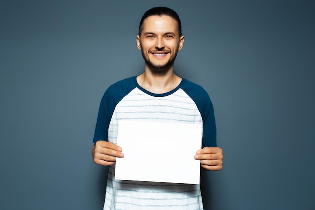 Foto retrato de estudio de un joven feliz con un tablero de papel blanco vacío en las manos sobre un fondo de color azul