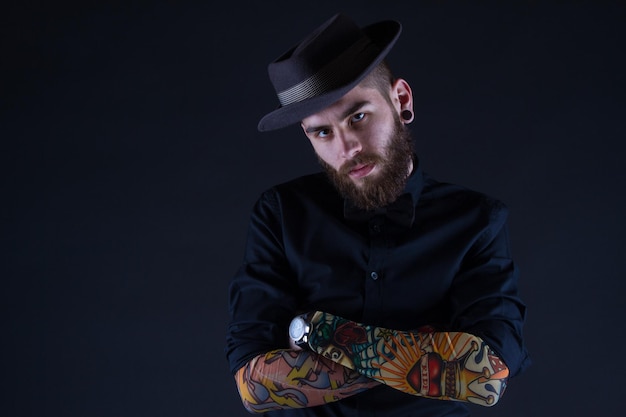 Retrato de estudio de un joven hipster de moda que usa elegantes mangas tatuadas y posa sobre un fondo negro