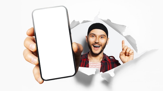 Retrato de estudio de un joven hombre feliz que muestra un gran teléfono inteligente con pantalla en blanco a través de un agujero de papel rasgado Fondo blanco Señalando con el dedo índice hacia arriba Nuevo concepto de idea