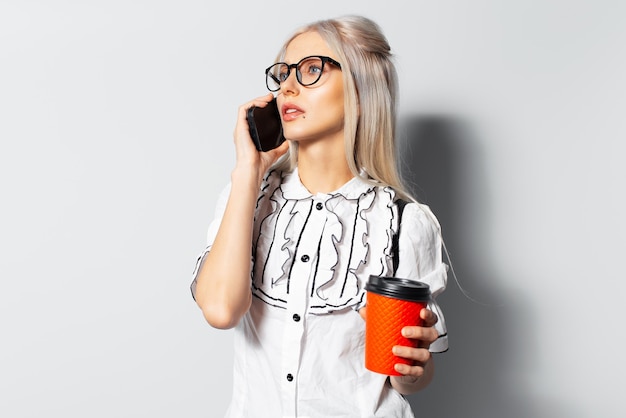 Retrato de estudio de una joven linda con cabello rubio usando un teléfono inteligente sosteniendo una taza de café para llevar usando anteojos sobre fondo blanco