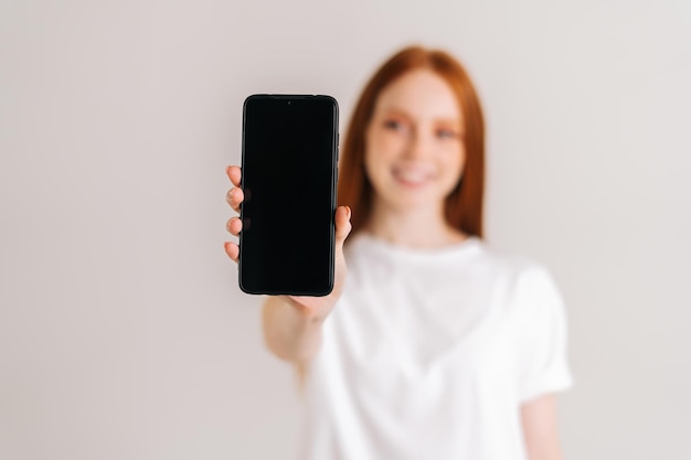 Retrato de estudio de una joven pelirroja positiva con una amplia sonrisa que muestra un teléfono móvil con pantalla en blanco