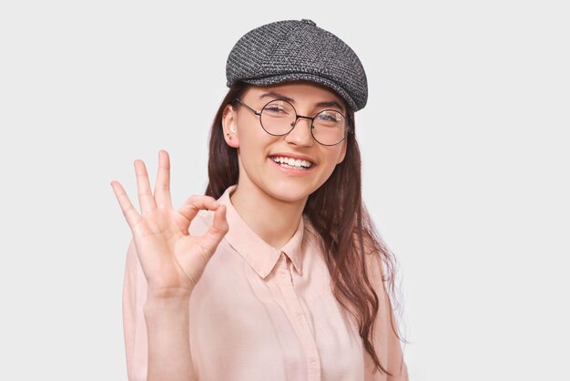 Retrato de estudio de una joven positiva que usa una camisa rosa de gorra gris de moda y anteojos redondos transparentes que muestran gestos de Ok Linda estudiante sonriendo parpadeando con un ojo y mostrando un signo de OK