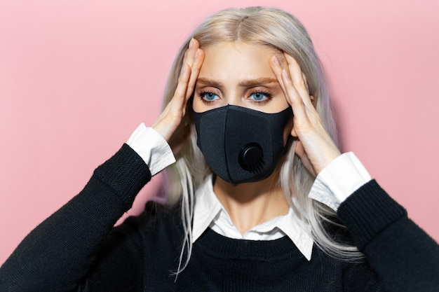 Retrato de estudio de una joven rubia cogidos de la mano en la cabeza debido a dolores, con máscara respiratoria contra el coronavirus sobre un fondo de color rosa pastel.