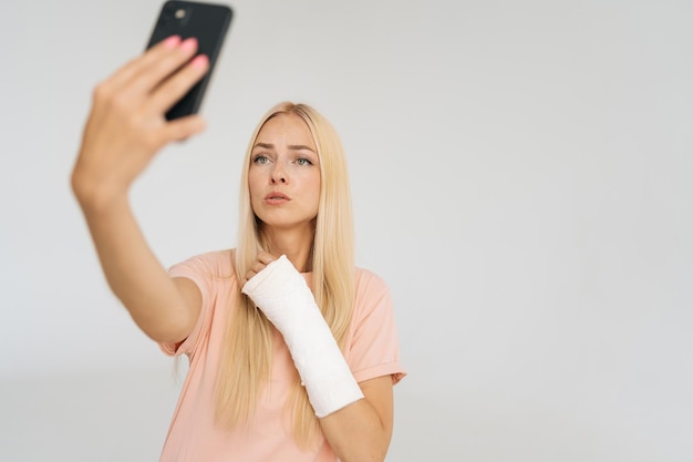 Retrato de estudio de una joven rubia seria con el brazo roto envuelto en una venda de yeso mirando a la pantalla del teléfono inteligente con la cara perpleja durante la videollamada