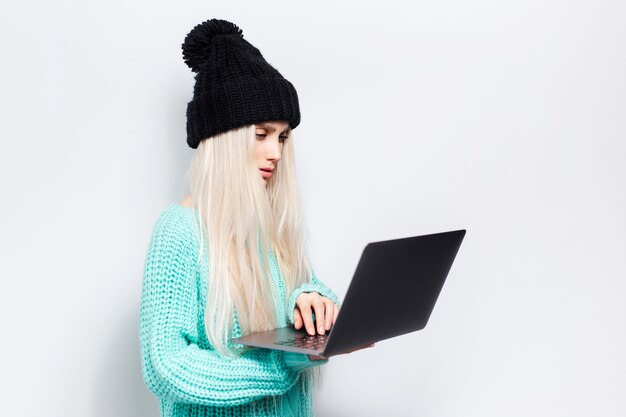 Retrato de estudio de joven rubia usando laptop sobre fondo blanco. Vistiendo sombrero negro y suéter cian.