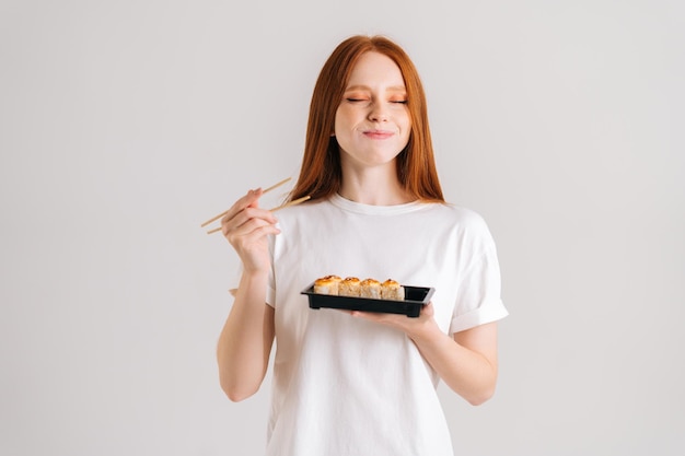 Retrato de estudio de una joven satisfecha con los ojos cerrados comiendo deliciosos rollos de sushi con