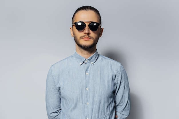 Retrato de estudio de un joven serio con gafas de sol y camisa azul de fondo blanco