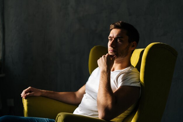 Retrato de estudio de un joven serio pensando en algo rascándose la barba sentado en un sillón amarillo en una habitación oscura